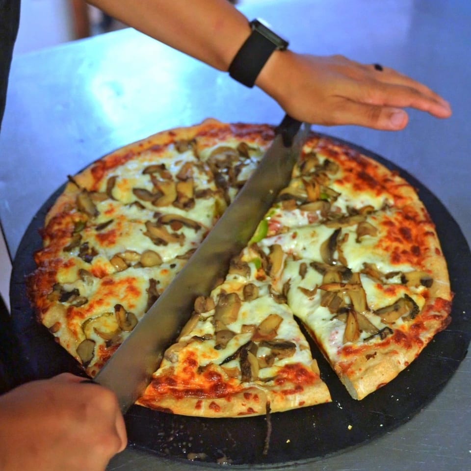 A hand slices a mushroom pizza on a black tray, with melted cheese and red sauce visible.